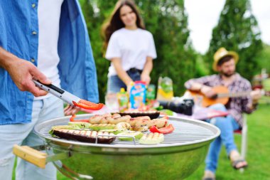 Erkeklerin elinde et ve sebzelerle ızgaranın arka planında lezzetli bir biftekle barbekü maşası ve piknikte eğlenen bir grup arkadaş var.
