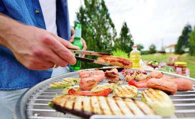 Mangalın arka planında sosis, sebze, kabak, biber, bira şişeleri, yeşillikler ve bir parkla ızgara sosisi olan bir adam elinde barbekü maşası tutuyor..