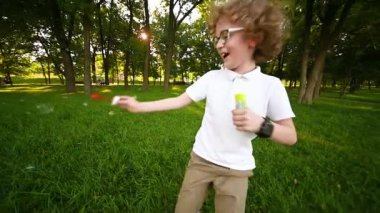 Gözlüklü, kıvırcık saçlı, sabun köpüğü üfleyerek yeşil çimlerin ve parkın arka planında koşan bir çocuk..