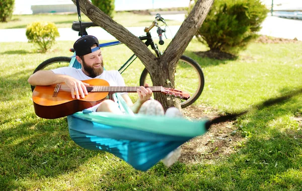 Γενειοφόρος άντρας με κιθάρα ξαπλωμένος σε μια αιώρα χαμογελώντας με φόντο τη φύση και το πράσινο. — Φωτογραφία Αρχείου
