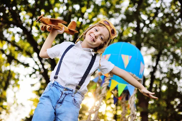 Маленький мальчик в белой рубашке с подтяжками с вьющимися волосами в шляпе пилота и очках играет с деревянным самолетом на фоне корзины с голубым шаром. — стоковое фото