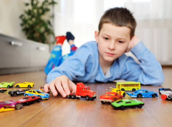 Preschool boy in a blue sweater is lying on the wooden floor playing with toy cars against the background of a bright room — Stock Photo, Image