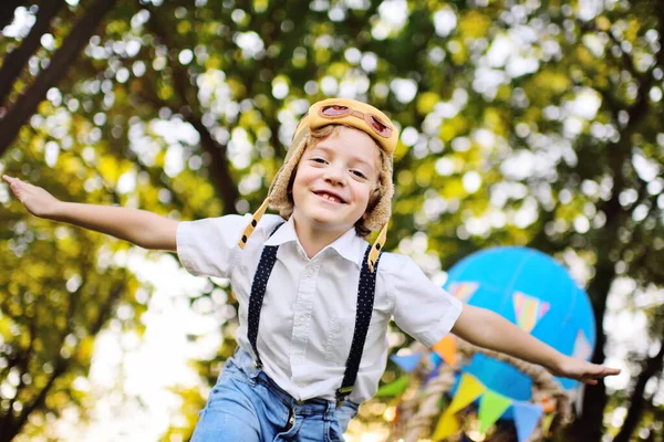 Boy in a white shirt with suspenders and wearing a pilots helmet with glasses smiles with his hands spread out depicting an airplane against a background of park and greenery — стоковое фото