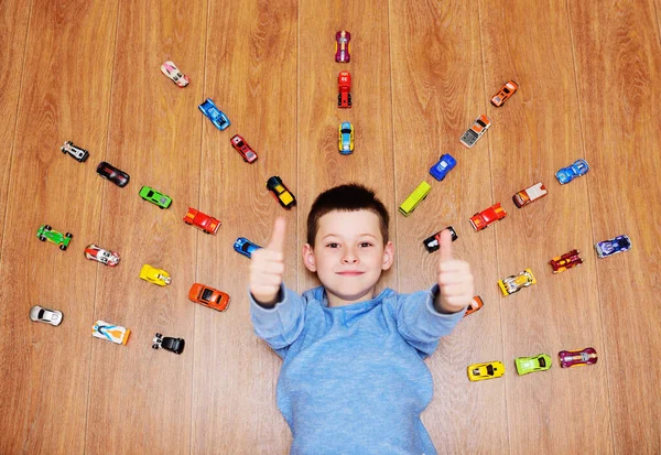 Klein kind een jongen in een blauwe trui ligt te lachen op de houten vloer tegen de achtergrond van veel speelgoed auto 's — Stockfoto