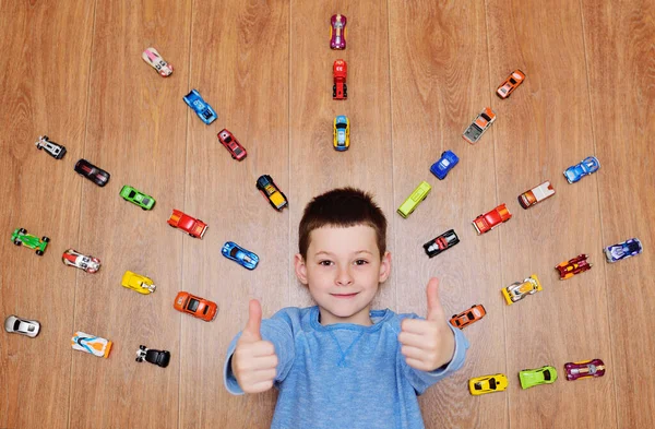 Klein kind een jongen in een blauwe trui ligt te lachen op de houten vloer tegen de achtergrond van veel speelgoed auto 's — Stockfoto