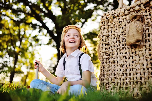 Small boy in a white shirt with suspenders and wearing a pilots helmet with glasses smiles as he sits on the grass leaning on a balloon basket a — стоковое фото