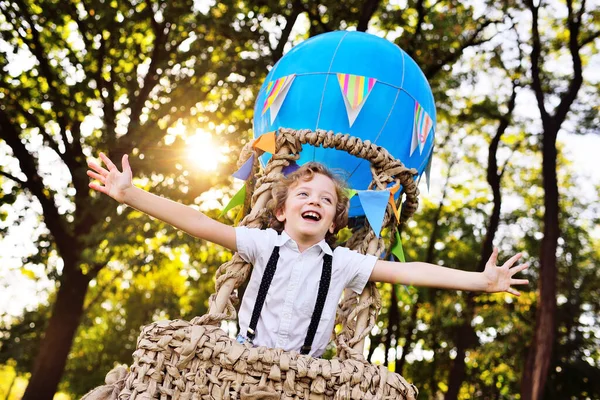 Мальчик с вьющимися волосами в корзине синего шарика улыбается и смотрит вдаль на фоне зелени и солнца. — стоковое фото