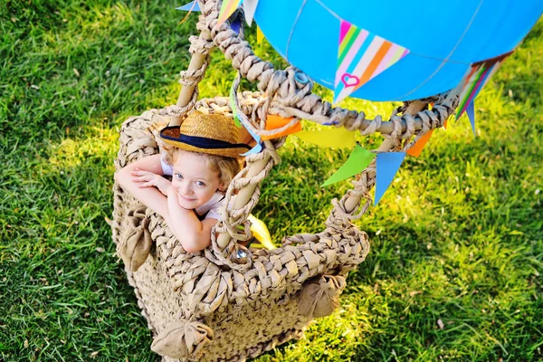 Mały chłopiec z kręconymi włosami w słomkowym kapeluszu siedzący w koszu z niebieskim balonem uśmiecha się na tle trawy — Zdjęcie stockowe