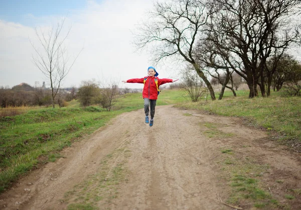 Pláza gyermek egy fiú piros kabátban és kék sapkában vidáman és boldogan ugrál és ugrál az úton a parkban vagy az erdőben. A gyermekkor fogalma, tavasz. — Stock Fotó