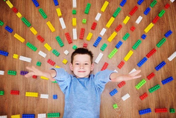 Een kleine jongen lacht tegen de achtergrond van kleurrijke blokken van kinderen bouw set met stralen op de vloer. — Stockfoto