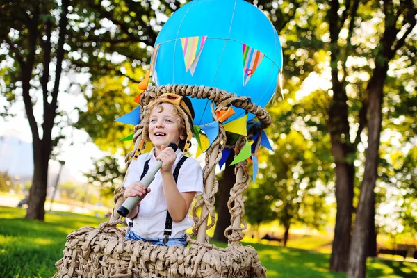 Small boy with curly hair in a pilots cap sitting with a telescope in his hands in a basket of a blue balloon smiles — стоковое фото