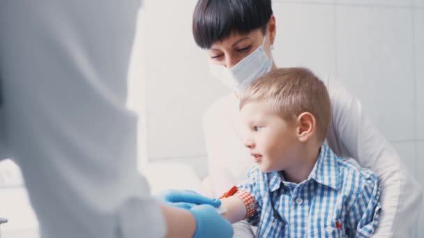 少年は分析のため静脈から血を取ってる。子供はクリニックの手術室で母親の腕の中に座っている. — ストック動画