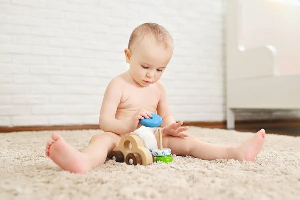 Cute baby sits on a carpet in a disposable diaper plays with toys against a white brick wall. — Stock Photo, Image