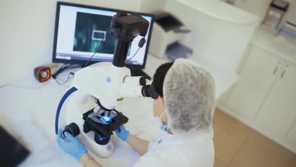 En läkare på en modern klinik arbetar bakom ett fluorescerande mikroskop. — Stockvideo