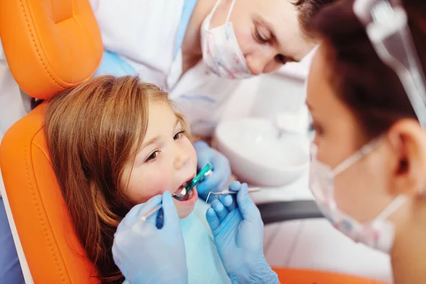 Dentistas jóvenes, un hombre y una mujer, examinan los dientes de un paciente infantil - una niña bonita que está sentada en una silla dental naranja. — Foto de Stock