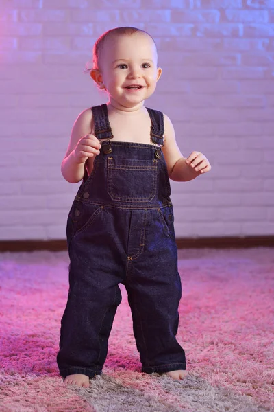 Baby boy in denim overalls smiling while standing barefoot on the carpet against the background of a white brick wall — Photo