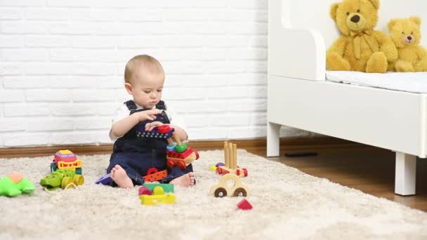 Baby boy in denim overalls plays with toys sitting on the carpet — Vídeos de Stock