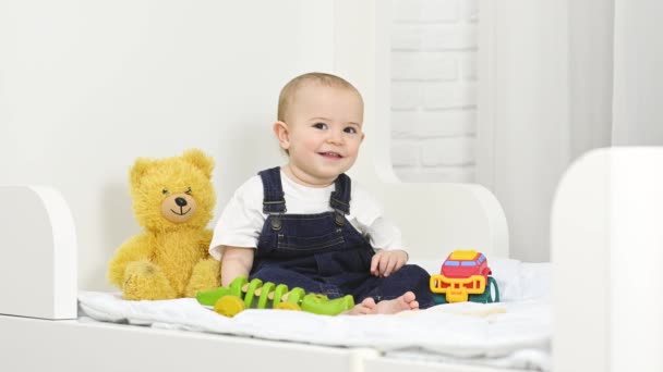 A baby boy in a denim jumpsuit smiles and plays with toys while sitting on a bed — Wideo stockowe