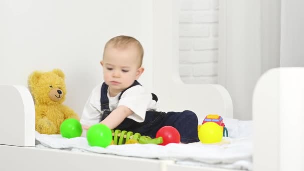 Bebé niño en overoles de mezclilla sentado en la cama juega juguetes y lanza bolas de plástico de color — Vídeo de stock