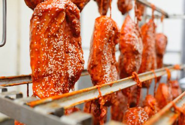 appetizing pieces of pork meat in a tomato marinade sprinkled with sesame seeds hang on a rack against the backdrop of a meat processing plant.
