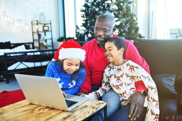 dad and two African American children make a video call through a laptop and communicate with relatives on New Years Eve against the background of the Christmas tree