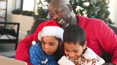 Afrika kökenli Amerikan ailesi - Noel Baba, oğul ve kız Noel Baba kazakları ve Noel Baba şapkalı kucaklaşma, gülümseme ve Noel ağacının arka planındaki akrabalarla video bağlantısı yoluyla iletişim.