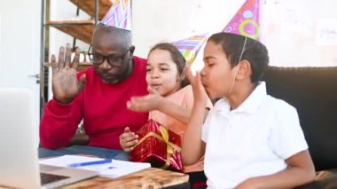 Afro-Amerikan ailesi - Baba, kız ve oğlan şenlikli şapkalarla doğum gününü kutluyor, gülümsüyor ve video bağlantısıyla akrabalarla iletişim kuruyor.