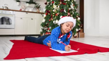 Mavi kazaklı ve şapkalı küçük bir Afro-Amerikalı Noel ağacının arkasına Noel Baba 'ya bir dilek mektubu yazıyor..