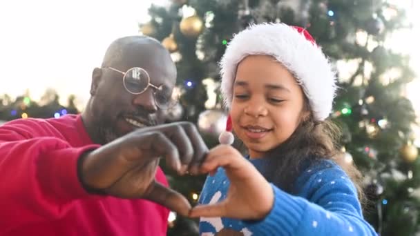 新年の衣装を着た父と小さなアフリカ系アメリカ人の娘は笑顔でクリスマスツリーの背景に手から心を作ります — ストック動画