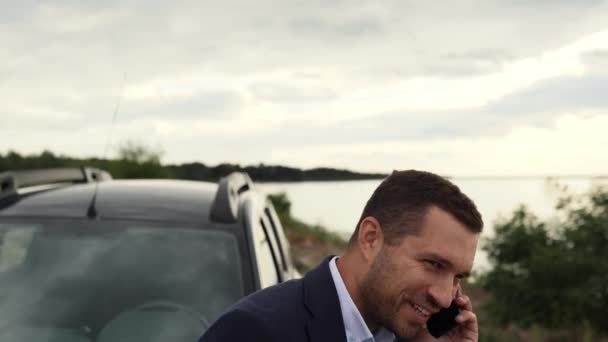 Un hombre de negocios está cerca de su coche en la costa de un lago, mar u océano, habla por teléfono, está feliz de nuevos éxitos en su negocio — Vídeos de Stock