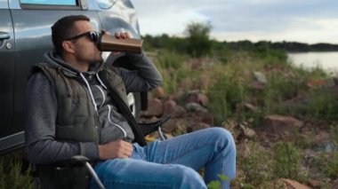 Bir adam göl, okyanus ya da deniz kıyısında bir araba gezisinde dinleniyor. Katlanan sandalyede bir koltuğu var. Elinde termos ya da termo bardak var.