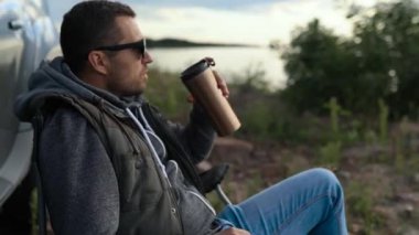 Bir adam göl, okyanus ya da deniz kıyısında bir araba gezisinde dinleniyor. Katlanan sandalyede bir koltuğu var. Elinde termos ya da termo bardak var.