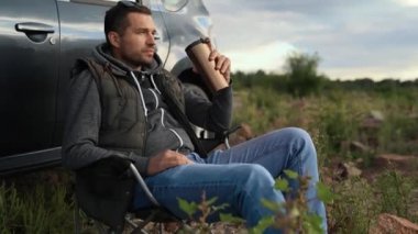 Bir adam göl, okyanus ya da deniz kıyısında bir araba gezisinde dinleniyor. Katlanan sandalyede bir koltuğu var. Elinde termos ya da termo bardak var.