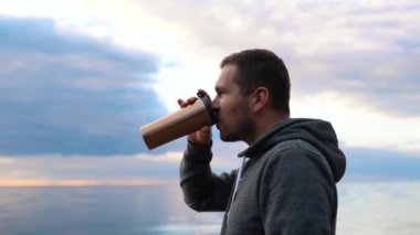 Sıcak bir termostan kahve ya da çay içen bir adam sakin bir denizde ya da okyanusta gün batımının arka planına karşı.