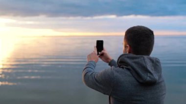 Bir adam gün batımını fotoğraflıyor ya da telefonda deniz kenarında video çekiyor.