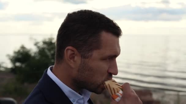 Un uomo d'affari si trova vicino alla sua auto sulla costa di un lago, mare o oceano, beve caffè e mangia un rotolo — Video Stock