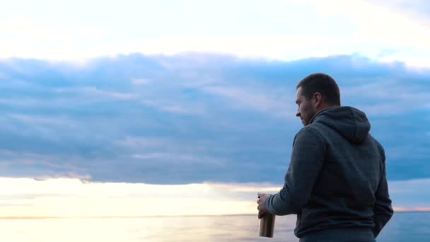 Un hombre que bebe café o té de un termo caliente sobre el telón de fondo de una puesta de sol en un mar o océano tranquilo — Vídeo de stock