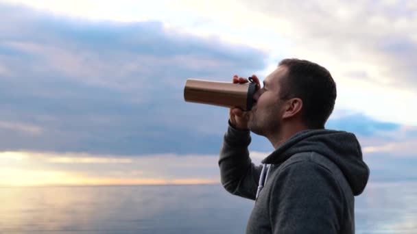 在平静的大海或大海中，在日落的背景下，在暖暖的热水瓶里喝咖啡或喝茶的人 — 图库视频影像