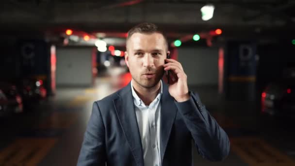 Businessman walks in the underground parking. He speaks on the phone and opens his car with the remote control — Stock Video