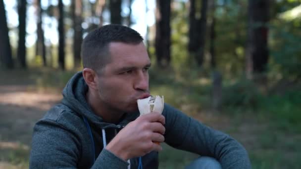 Mężczyzna w parku lub lesie na poboczu siada, zjada bułkę i pije wodę z plastikowej butelki po ciężkim treningu ulicznym. — Wideo stockowe