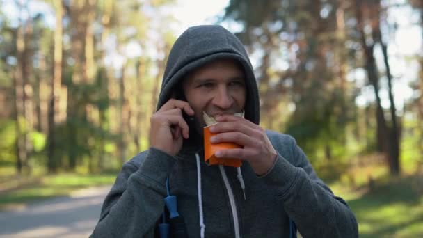 Ein Mann telefoniert und isst nach einem harten Training ein Brötchen im Wald oder Park. Ein Springseil pfeift um seinen Hals. — Stockvideo
