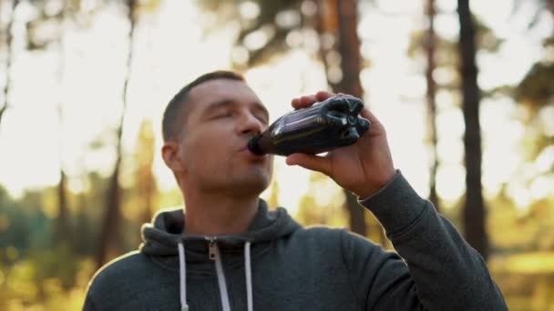 Egy férfi az erdőben vagy a parkban iszik vizet egy műanyag hulladékból egy kemény kocogás edzés után — Stock videók