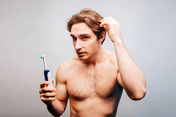 El hombre sostiene el cepillo de dientes eléctrico y toca su cabello Imágenes De Stock Sin Royalties Gratis