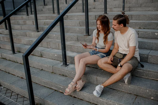 Encantadora pareja está sentada en las escaleras afuera y mirando en el teléfono inteligente — Foto de Stock