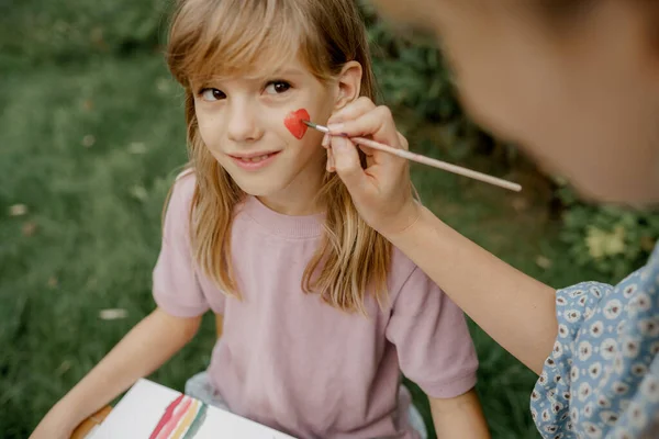 Giovane madre sta disegnando da vernici sul suo volto bambino in giardino Immagini Stock Royalty Free