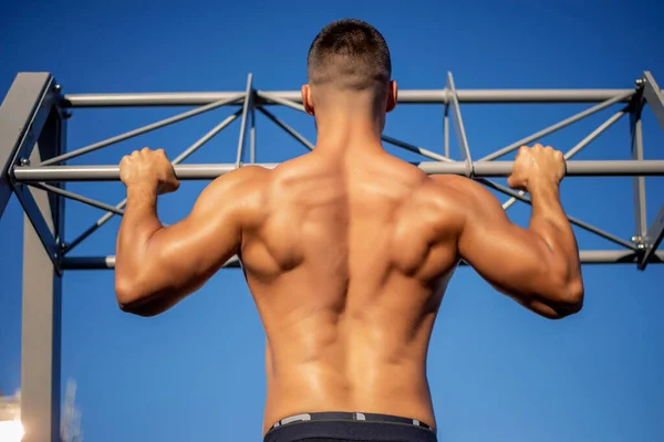 Back view of muscular sportsman doing pull ups on horizontal bar at gym. Sports trainer in gym workout exercising. Fit and athletic man having naked torso, wearing in black shorts.