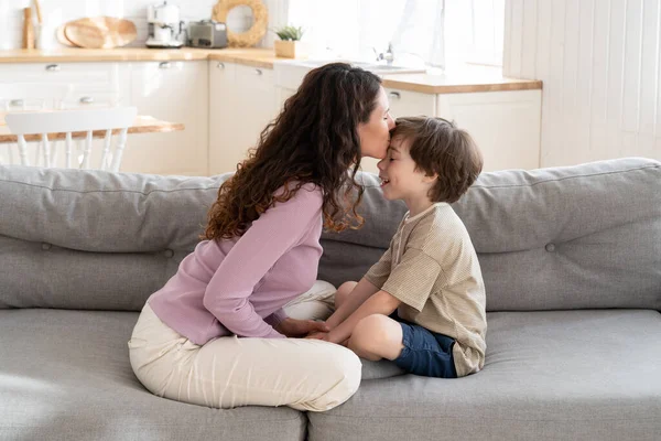 Šťastná rodina: milující maminka líbání malého syna na čele sedí se zkříženýma nohama na pohovce doma — Stock fotografie