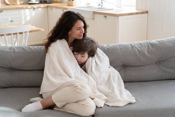 Mladá máma sedět na útulném gauči relaxovat s předškolním synem pokryté dekou o víkendu ráno doma — Stock fotografie