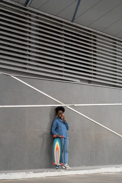 Африканська дівчина-ковзаняр робить телефонний дзвінок, тримаючи смартфон з довгою дошкою біля стіни будівлі. — стокове фото