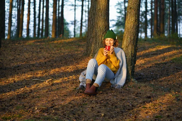Отдых на природе: счастливая женщина сидит под деревом в осеннем лесу и пьет горячий чай, наслаждаясь закатом — стоковое фото